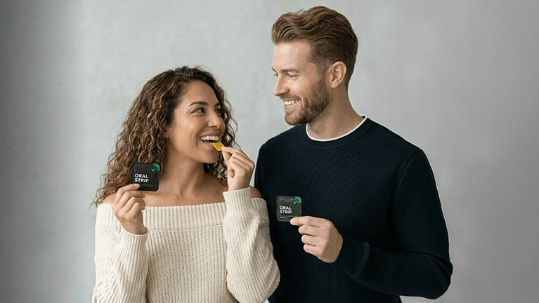 Man and woman holding Ora2M toothpaste tubes against a gray background
